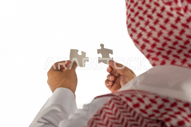 بورتريه بصورة مقربة ليد رجل سعودي خليجي يحمل قطعتين من اللغز يحاول ربط وتوصيل قطع اللغز الصغيرة ، مفهوم التركيز وحل اللغز ، التسليه والترفيه ، لعبة بازل ، لعبة تركيب الصور على خلفية بيضاء