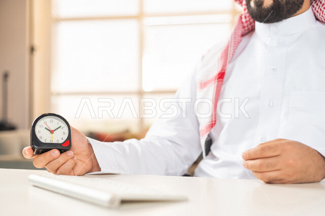 صورة مقربة لرجل أعمال سعودي خليجي يمسك ساعة رقمية صغيرة ، استخدام لوحة المفاتيح للكتابة ، مفهوم إدارة وقت العمل ، الدقة في المواعيد ، التذكيرات والتنبيهات ، العمل عن بعد ، التسويق الإلكتروني ، إنجاز المهامات في الوقت