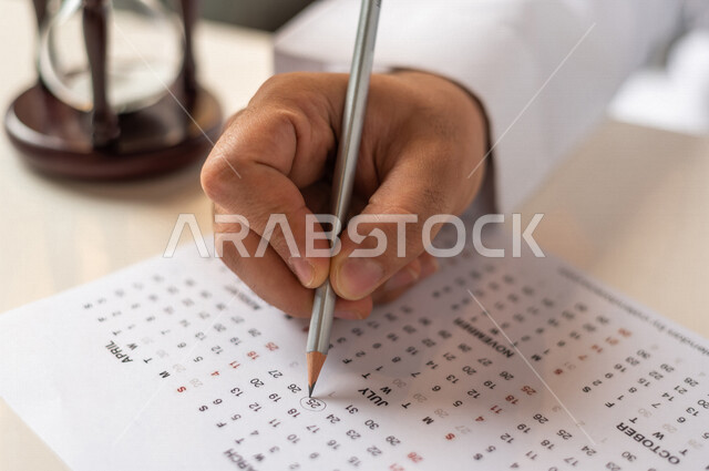 صورة مقربة ليد رجل سعودي خليجي يستخدم القلم لجدول الكتابة في التقويم، الساعة الرملية الزجاجية (البنكام) على الطاولة ، مفهوم إدارة الأعمال والوقت، توقيت الرمل، تحديد مواعيد العمل ، مفهوم إدارة الجدول الزمني ، قياس الوقت