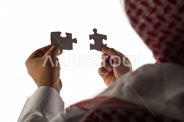 بورتريه بصورة مقربة ليد رجل سعودي خليجي يحمل قطعتين من اللغز يحاول ربط وتوصيل قطع اللغز الصغيرة ، مفهوم التركيز وحل اللغز ، التسليه والترفيه ، لعبة بازل ، لعبة تركيب الصور على خلفية بيضاء