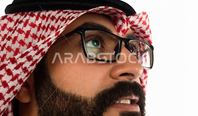 بورتريه بصورة مقربة لرجل سعودي خليجي يقوم بجلسة تصوير ، ينظر لأعلى ، خلفية بيضاء لرجل سعودي مسلم يرتدي زي تقليدي سعودي مع شماغ وعقال