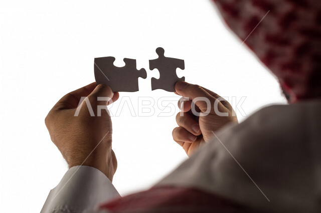 بورتريه بصورة مقربة ليد رجل سعودي خليجي يحمل قطعتين من اللغز يحاول ربط وتوصيل قطع اللغز الصغيرة ، مفهوم التركيز وحل اللغز ، التسليه والترفيه ، لعبة بازل ، لعبة تركيب الصور على خلفية بيضاء