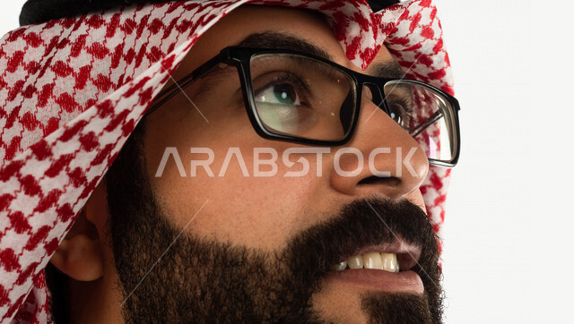 بورتريه بصورة مقربة لرجل سعودي خليجي يقوم بجلسة تصوير ، ينظر لأعلى ، خلفية بيضاء لرجل سعودي مسلم يرتدي زي تقليدي سعودي مع شماغ وعقال