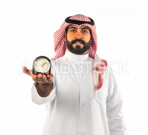 Portrait of a Saudi Gulf man holding a small digital watch with different face and hands gestures, white background of a Saudi man wearing traditional Saudi dress with shemagh and iqal, concept of working time management, punctuality, constant reminders a