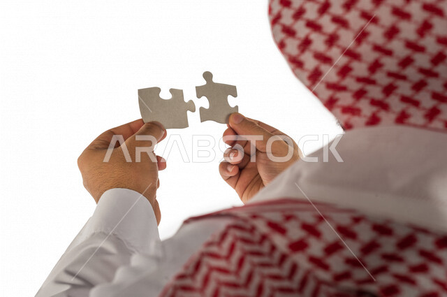 بورتريه بصورة مقربة ليد رجل سعودي خليجي يحمل قطعتين من اللغز يحاول ربط وتوصيل قطع اللغز الصغيرة ، مفهوم التركيز وحل اللغز ، التسليه والترفيه ، لعبة بازل ، لعبة تركيب الصور على خلفية بيضاء