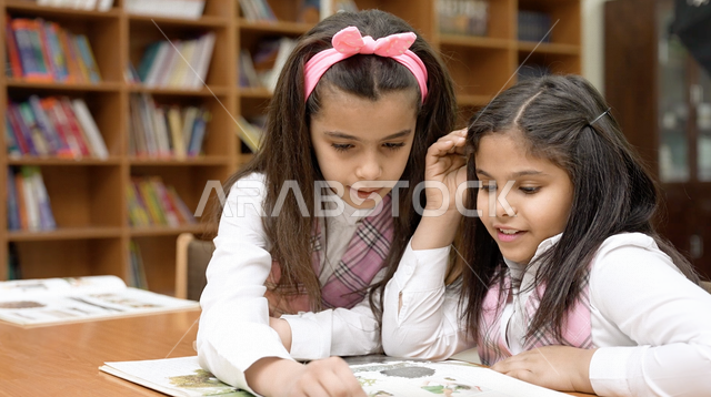 صورة لتلميذتان عربيتان خليجيتان سعوديتان تجلسان في المكتبة المدرسية، الاستمتاع بقراءة الكتب المدرسية، مكتبة المدرسة، تأدية الواجبات المدرسية، التعليم في المملكة العربية السعودية