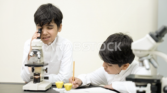 تلميذان عربيان خليجيان سعوديان يرتديان الزي المدرسي، اجراء البحوثات والاختبارات باستخدم التلسكوب داخل مختبر المدرسة، بحوث علمية، تسجيل الملاحظات، التعليم في المملكة العربية السعودية