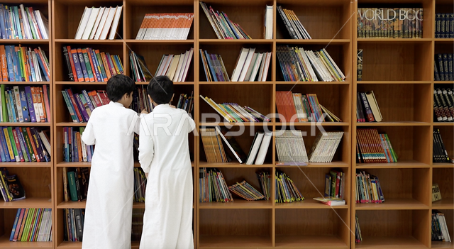 صورة من الخلف لتلميذان عربيان خليجيان سعوديان يقفان امام رفوف الكتب في المكتبة المدرسية، البحث عن كتب مدرسية، مكتبة المدرسة، التعليم في المملكة العربية السعودية