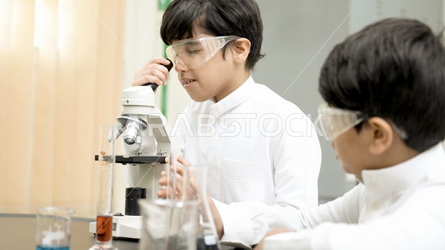 تلميذان عربيان خليجيان سعوديان يرتديان الزي المدرسي، اجراء البحوثات والاختبارات باستخدم التلسكوب داخل مختبر المدرسة، بحوث علمية، تسجيل الملاحظات، التعليم في المملكة العربية السعودية