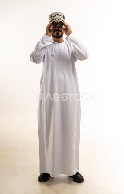 إتقان واحتراف مهنة التصوير، بورتريه لشاب عربي خليجي عماني مبتسم يرتدي يرتدي الدشداشة و الكمه يقوم بالتقاط صور تذكارية عبر كاميرا فوتوغرافية، تعابير الفرح والسرور، استخدام كاميرات رقمية عالية الدقة، وظائف ومهن سعودية، صورة الجسم بالطول الكامل، خلفية بيضاء