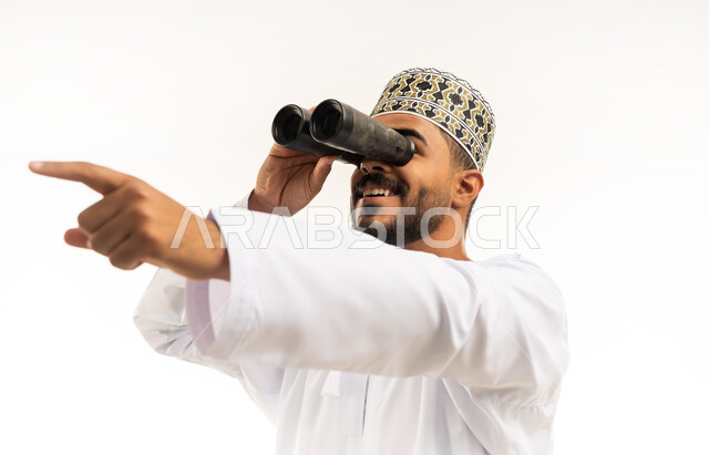 رؤية الاشياء البعيدة عبر المنظار، حب المغامرة والاستكشاف، بورتريه مقرب لشاب عربي خليجي عماني يرتدي الدشداشة والكمه ينظر من خلال المنظور ثنائي العينين يشير بإصبع السبابة الى شيء ما بإيماءات الفرح والسرور، خلفية بيضاء