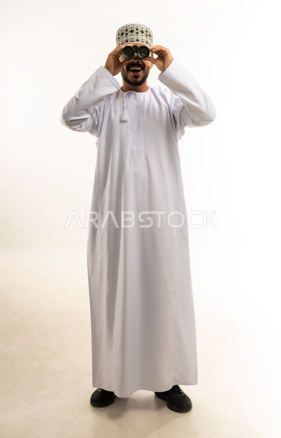 ايماءات الاستمتاع و السرور, رؤية الاشياء البعيدة و تقريبها, حب الاستكشاف و المغامرة, بورتريه لشاب عربي خليجي عماني يرتدي الدشداشة و الكمه مبتسم يحمل بيده منظار ثنائي العينين و يينظر الى شيئ ما, صورة الجسم بالطول الكامل, خلفية بيضاء