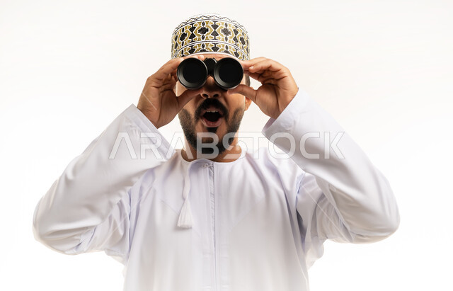 ايماءات المفاجئة والتعجب, رؤية الاشياء البعيدة و تقريبها, حب الاستكشاف و المغامرة, بورتريه مقرب لشاب عربي خليجي عماني يرتدي الدشداشة و الكمه مبتسم يحمل بيده منظار ثنائي العينين و يينظر الى شيئ ما, خلفية بيضاء