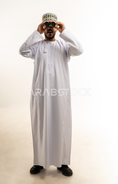 رؤية الاشياء البعيدة و تقريبها, حب الاستكشاف و المغامرة, ايماءات المفاجئة والتعجب, بورتريه لشاب عربي خليجي عماني يرتدي الدشداشة و الكمه مبتسم يحمل بيده منظار ثنائي العينين و يينظر الى شيئ ما, صورة الجسم بالطول الكامل, خلفية بيضاء
