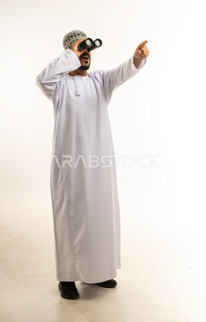 حب المغامرة والاستكشاف، رؤية الاشياء البعيدة عبر المنظار، بورتريه لشاب عربي خليجي عماني يرتدي الدشداشة والكمه ينظر من خلال المنظور ثنائي العينين يشير بإصبع السبابة الى شيء ما بإيماءات الفرح والسرور، صورة الجسم بالطول الكامل، خلفية بيضاء