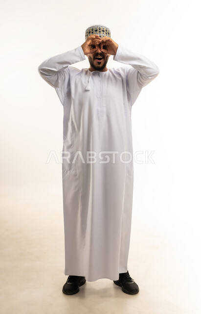 ايماءات الاستمتاع و السرور, رؤية الاشياء البعيدة و تقريبها, حب الاستكشاف و المغامرة, بورتريه لشاب عربي خليجي عماني يرتدي الدشداشة و الكمه مبتسم  يضع يديه على عينه على شكل المنظار ينظر الى شيئ ما, صورة الجسم بالطول الكامل, خلفية بيضاء