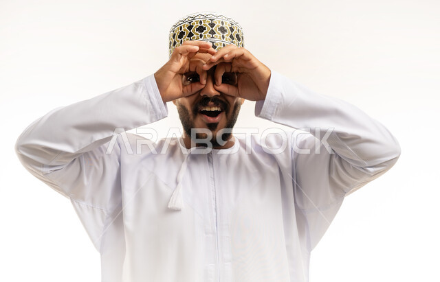 رؤية الاشياء البعيدة و تقريبها, ايماءات الاستمتاع و السرور, حب الاستكشاف و المغامرة, بورتريه مقرب لشاب عربي خليجي عماني يرتدي الدشداشة و الكمه مبتسم  يضع يديه على عينه على شكل المنظار ينظر الى شيئ ما, خلفية بيضاء