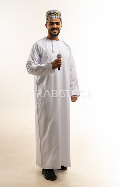 العمل في تقديم الفقرات و البرامج الفنية الترفيهية, النظر الى الكاميرا بإيماءات السعادة, بورتريه لشاب عربي خليجي عماني يرتدي الدشداشة و الكمة يمسك بيده المايكروفون، الجسم بالطول الكامل, خلفية بيضاء