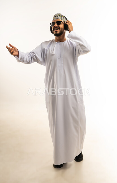 التفاعل والاندماج مع الأنغام الموسيقية، مواهب غنائية فنية, الاستماع للأغاني باسخدام الهيدفون، بورتريه لشاب عربي خليجي عماني يرتدي الدشداشة والكمه يضع سماعات الرأس بإيماءات الفرح والسرور، صورة الجسم بالطول الكامل, خلفية بيضاء