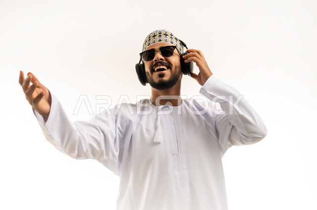 مواهب غنائية فنية, الاستماع للأغاني باسخدام الهيدفون، التفاعل والاندماج مع الأنغام الموسيقية، بورتريه مقرب لشاب عربي خليجي عماني يرتدي الدشداشة والكمه يضع سماعات الرأس بإيماءات الفرح والسرور، خلفية بيضاء