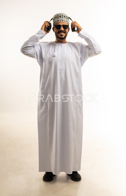 التفاعل والاندماج مع الأنغام الموسيقية، مواهب غنائية فنية, الاستماع للأغاني باسخدام الهيدفون، بورتريه لشاب عربي خليجي عماني يرتدي الدشداشة والكمه يضع سماعات الرأس بإيماءات الفرح والسرور، صورة الجسم بالطول الكامل, خلفية بيضاء