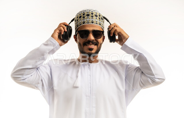 مواهب غنائية فنية, الاستماع للأغاني باسخدام الهيدفون، التفاعل والاندماج مع الأنغام الموسيقية، بورتريه مقرب لشاب عربي خليجي عماني يرتدي الدشداشة والكمه يضع سماعات الرأس بإيماءات الفرح والسرور، خلفية بيضاء