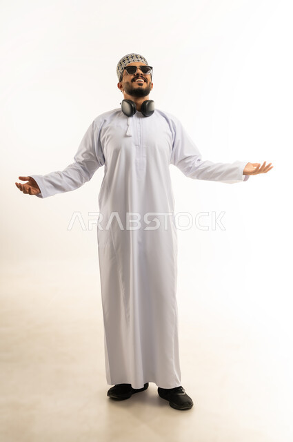 The concept of achievement and goal attainment, victory, winning and success, portrait of a smiling young Arab Gulf Omani man wearing a dishdasha and a cloak, looking at the camera, raising his hands up with gestures of joy and pleasure, feeling happy and strong, full-length body image, white background