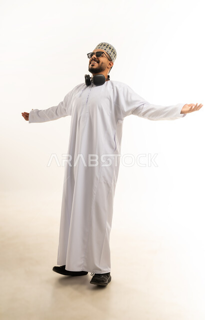 الشعور بالسعادة والقوة, مفهوم الانجاز وتحقيق الاهداف، الانتصار والفوز والنجاح، بورتريه لشاب عربي خليجي عماني مبتسم يرتدي الدشداشة و الكمه ينظر الى الكاميرا يرفع يديه للاعلى بإيماءات الفرح والسرور, صورة الجسم بالطول الكامل, خلفية بيضاء