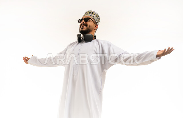 الانتصار والفوز والنجاح، الشعور بالسعادة والقوة, مفهوم الانجاز وتحقيق الاهداف، بورتريه لشاب عربي خليجي عماني مبتسم يرتدي الدشداشة و الكمه ينظر الى الكاميرا يرفع يديه للاعلى بإيماءات الفرح والسرور, خلفية بيضاء