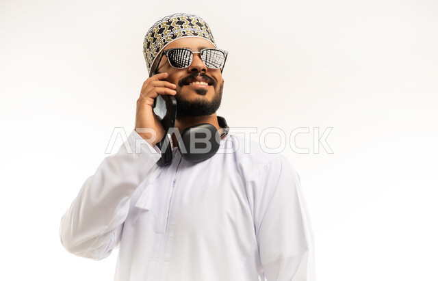 التواصل مع الاهل و الاصدقاء, ايماءات السعادة و السرور، استخدام التقنيات الحديثة المتطورة, بورتريه مقرب لشاب عربي خليجي عماني يرتدي الدشداشه و الكمه يقوم بإجراء مكالمة هاتفية عبر الهاتف المحمول، خلفية بيضاء