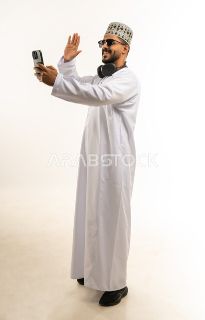 رفع كف اليد لالقاء التحية ورد السلام, إجراء مكالمة فيديو باستخدام الموبايل، تبادل الأخبار مع الأهل والأصدقاء، بورتريه لشاب عربي خليجي عماني يرتدي الدشداشة والكمه يقوم بالتواصل مع الأقارب، تعابير الفرح والسرور، صورة الجسم بالطول الكامل, خلفية بيضاء
