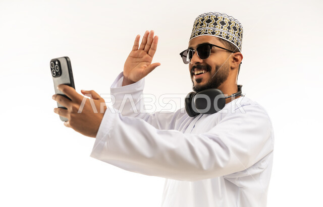 إجراء مكالمة فيديو باستخدام الموبايل، رفع كف اليد لالقاء التحية ورد السلام, تبادل الأخبار مع الأهل والأصدقاء، بورتريه مقرب لشاب عربي خليجي عماني يرتدي الدشداشة والكمه يقوم بالتواصل مع الأقارب، تعابير الفرح والسرور، خلفية بيضاء