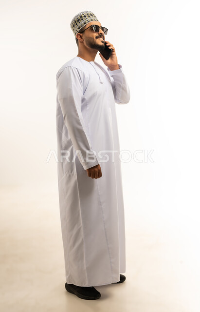 Using advanced modern technologies, gestures of happiness and pleasure, portrait of a young Arab Gulf Omani man wearing a dishdasha and a turban making a phone call via a mobile phone, communicating with family and friends, full-length body image, white background