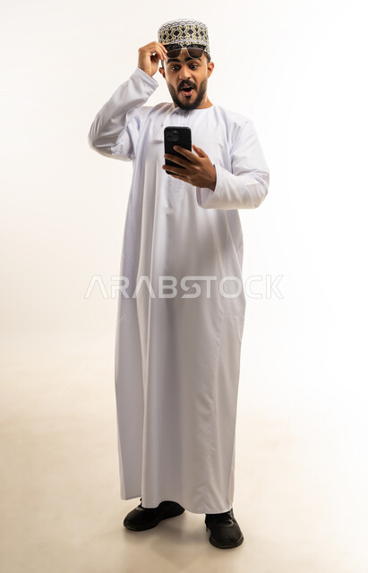 مشاهدة الأخبار المفاجئة والصادمة عبر الجوال, تعابير الاستغراب والاندهاش, استخدام التقنيات الحديثة المتطورة, بورتريه لشاب عربي خليجي عماني يرتدي الدشداشه و الكمه يقوم باستخدام الهاتف المحمول, صورة الجسم بالطول الكامل, خلفية بيضاء