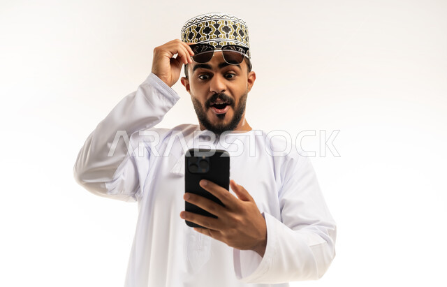 تعابير الاستغراب والاندهاش, مشاهدة الأخبار المفاجئة والصادمة عبر الجوال, استخدام التقنيات الحديثة المتطورة, بورتريه مقرب لشاب عربي خليجي عماني يرتدي الدشداشه و الكمه يقوم باستخدام الهاتف المحمول, خلفية بيضاء