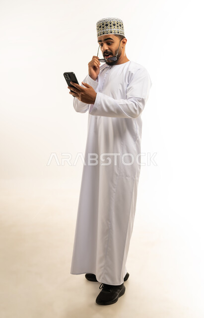 إمكانية تسديد الفواتير عبر بطاقة الصراف, ايماءات المفاجئة والتعجب, التقنيات الحديثة المتطورة، استخدام تطبيقات الجوال للدفع والشراء، بورتريه لشاب عربي خليجي عماني يرتدي الدشداشة و الكمة يمسك بيده البطاقة البنك ينظر الى الهاتف, الجسم بالطول الكامل, خلفية بيضاء