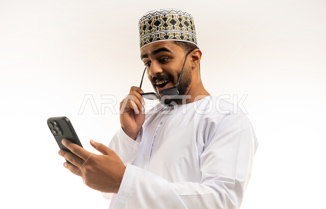 ايماءات المفاجئة والتعجب, التقنيات الحديثة المتطورة، استخدام تطبيقات الجوال للدفع والشراء، إمكانية تسديد الفواتير عبر بطاقة الصراف, بورتريه مقرب لشاب عربي خليجي عماني مبتسم يرتدي الدشداشة و الكمة يمسك بيده البطاقة الالكترونية ينظر الى الهاتف المحمول, خلفية بيضاء