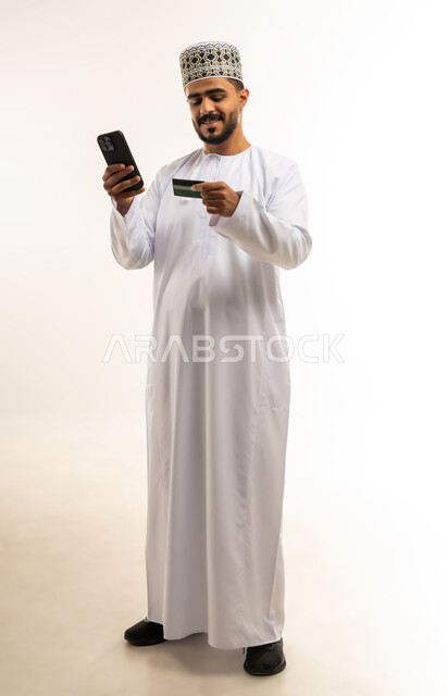 ايماءات الفرح والسعادة, التقنيات الحديثة المتطورة، استخدام تطبيقات الجوال للدفع والشراء، إمكانية تسديد الفواتير عبر بطاقة الصراف, بورتريه لشاب عربي خليجي عماني مبتسم يرتدي الدشداشة و الكمة يمسك بيده البطاقة الالكترونية ينظر الى الهاتف المحمول, صورة الجسم بالطول الكامل, خلفية بيضاء