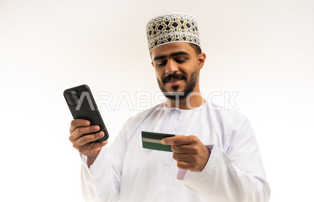 Modern advanced technologies, use of mobile applications for payment and purchase, possibility of paying bills via debit card, close-up portrait of a smiling young Arab Gulf Omani man wearing a dishdasha and a kummah holding an electronic card in his hand, looking at the camera with gestures of pleasure, white background