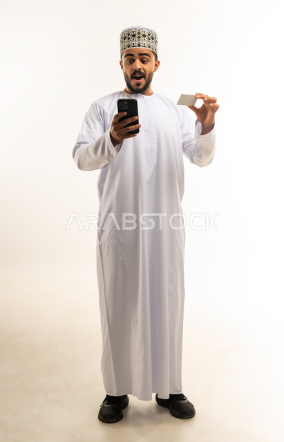 إمكانية تسديد الفواتير عبر بطاقة الصراف, استخدام تطبيقات الجوال للدفع والشراء، التقنيات الحديثة المتطورة، بورتريه لشاب عربي خليجي عماني مبتسم يرتدي الدشداشة و الكمة يمسك بيده البطاقة الالكترونية ينظر الى الكاميرا بإيماءات المفاجئة, صورة الجسم بالطول الكامل, خلفية بيضاء