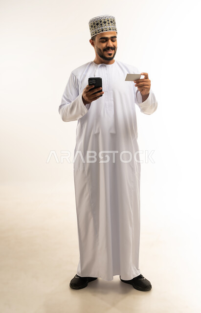 استخدام تطبيقات الجوال للدفع والشراء، إمكانية تسديد الفواتير عبر بطاقة الصراف, التقنيات الحديثة المتطورة، بورتريه لشاب عربي خليجي عماني مبتسم يرتدي الدشداشة و الكمة يمسك بيده البطاقة الالكترونية ينظر الى الكاميرا بإيماءات السرور, صورة الجسم بالطول الكامل, خلفية بيضاء
