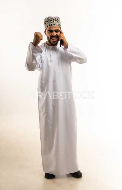 اتصالات ودردشة مع الأهل والأصدقاء, بورتريه لشاب عربي خليجي عماني مبتسم يرتدي الدشداشة و الكمه يمسك بيده الهاتف المحمول يقوم بإجراء مكالمة هاتفية يرفع قبضة يده ينظر الى الكاميرا بإيماءات الفوز والانتصار, سماع الاخبار السارة، صورة الجسم بالطول الكامل, خلفية بيضاء