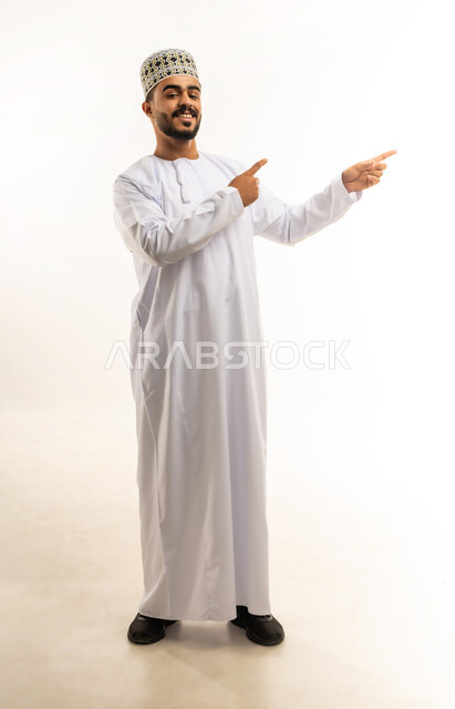 تحديد موقع معين, الإشارة و التوضيح إلى شيء ما, الدلالة على مكان عنصر، بورتريه لشاب عربي خليجي عماني مبتسم يرتدي الدشداشة و الكمه ينظر الى الكاميرا يشير بإصبع السبابة إلى شيء ما بإيماءات السرور, صورة الجسم بالطول الكامل، خلفية بيضاء
