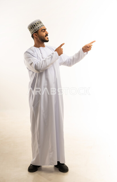 الإشارة و التوضيح إلى شيء ما, تحديد موقع معين, الدلالة على مكان عنصر، بورتريه لشاب عربي خليجي عماني مبتسم يرتدي الدشداشة و الكمه يشير بإصبع السبابة إلى شيء ما ينظر اليه بإيماءات السرور, صورة الجسم بالطول الكامل، خلفية بيضاء