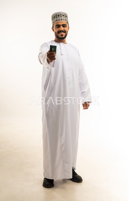 Possibility of paying bills via debit card, purchase and payment via bank credit cards, close-up portrait of a smiling young Arab Gulf Omani man wearing casual clothes holding an electronic card in his hand looking at the camera with gestures of pleasure, full-length body image, white background