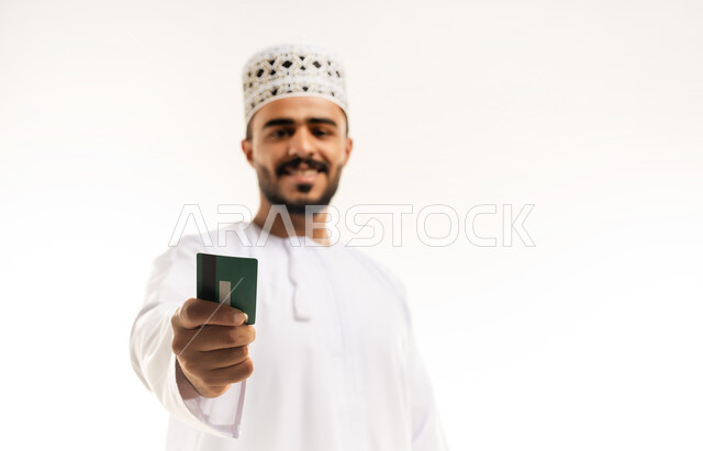 الشراء والدفع عن طريق بطاقات الإئتمان البنكية، إمكانية تسديد الفواتير عبر بطاقة الصراف, بورتريه مقرب مقرب لشاب عربي خليجي عماني مبتسم يرتدي الزي الكاجوال يمسك بيده البطاقة الالكترونية ينظر الى الكاميرا بإيماءات السرور, خلفية بيضاء