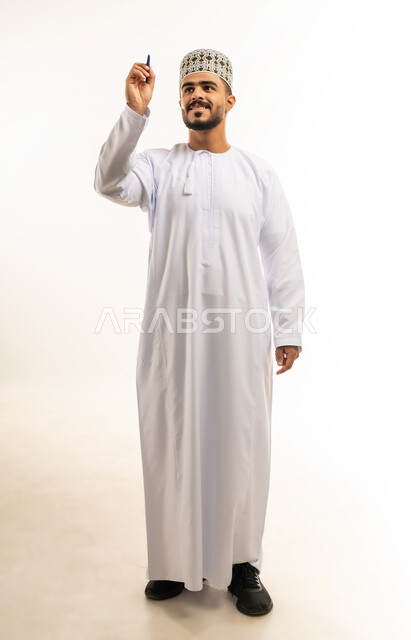 Taking notes, preparing to write and specify something, portrait of a young Arab Gulf Omani man wearing a dishdasha and a cloak holding a pen writing something, concept of education according to the prescribed curricula, full-length body image, white background