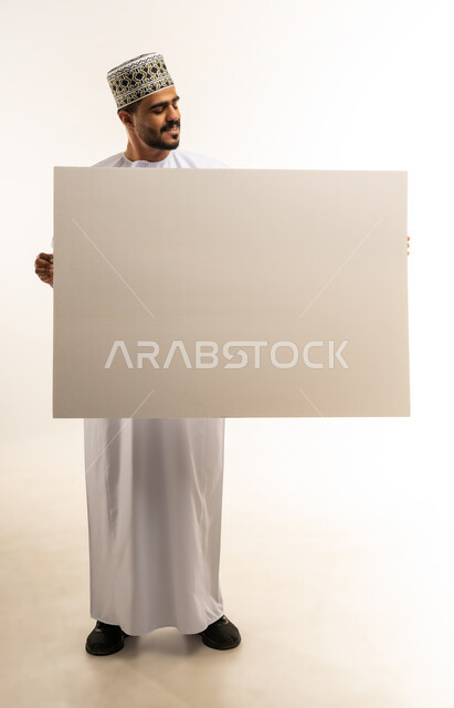 إيماءات الفرح و السرور، إعلانات وعروضات تسويقية، استخدام لوحات توضيحية تعريفية، موك اب مستطيل أبيض، بورتريه لشاب عربي خليجي عماني مبتسم يرتدي الدشداشة والكمه يحمل بيده لوحه بيضاء مستطيلة كبيرة فارغة, صورة الجسم بالطول الكامل, خلفية بيضاء
