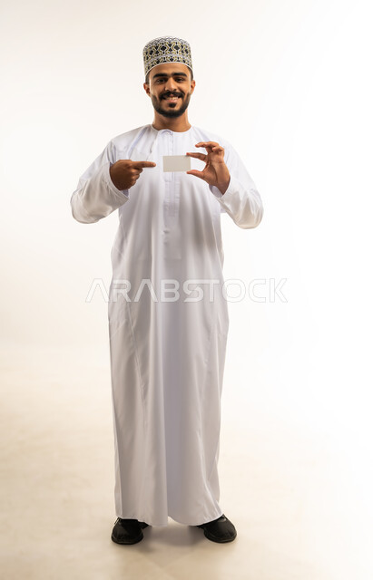 Using identification panels, a small white rectangular mockup, a portrait of a smiling young Omani Gulf Arab man wearing a dishdasha and a sleeve holding an empty white rectangular board looking at the camera with gestures of joy and happiness, advertisements and marketing offers, a full-length body image, a white background