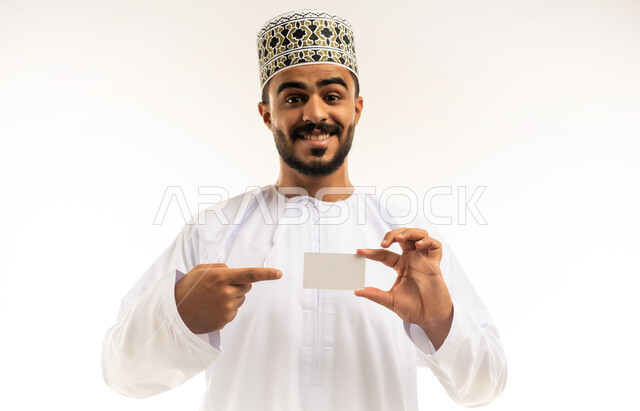 إعلانات وعروضات تسويقية، استخدام لوحات توضيحية تعريفية، موك اب مستطيل أبيض صغير، بورتريه لشاب عربي خليجي عماني مبتسم يرتدي الدشداشة والكمه يحمل بيده لوحه بيضاء مستطيلة فارغة ينظر الى الكاميرا بإيماءات الفرح السرور، خلفية بيضاء